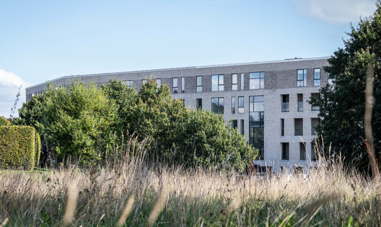 Moderne lejlighedsbyggeri omgivet af grønt græs og træer under blå himmel.