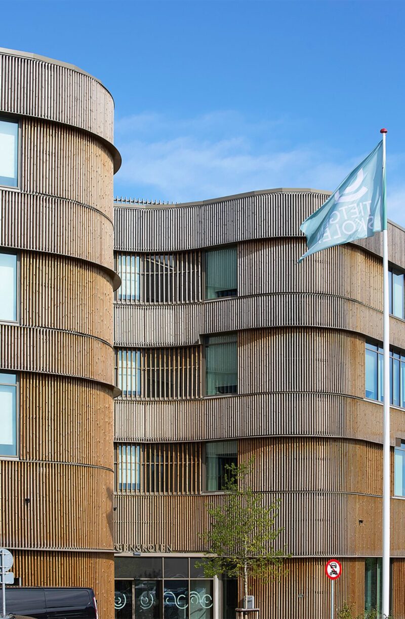 Moderne bygning med træpaneler og vinduer, blå himmel i baggrunden, flagstang med flag ses i forgrunden.