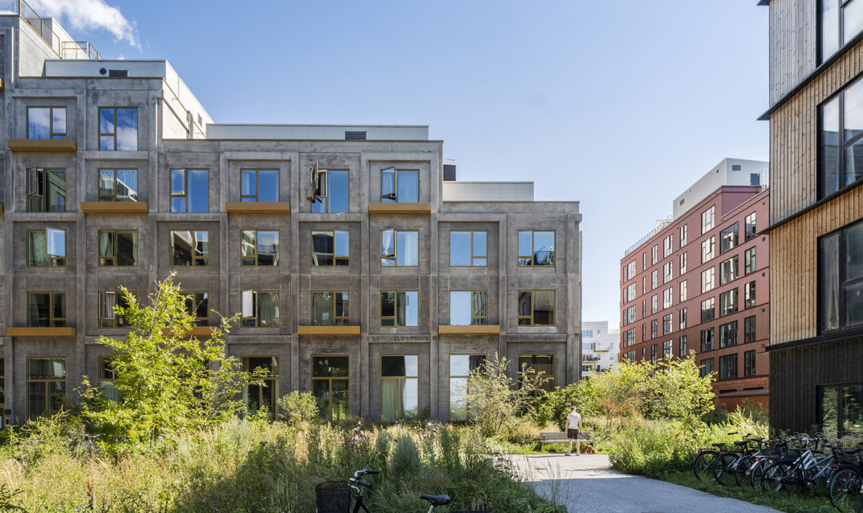 Moderne boligbyggeri med grå betonfacade, grønne områder og cykler i et bymiljø under blå himmel.