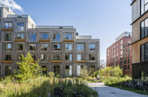 Moderne boligbyggeri med grå betonfacade, grønne områder og cykler i et bymiljø under blå himmel.