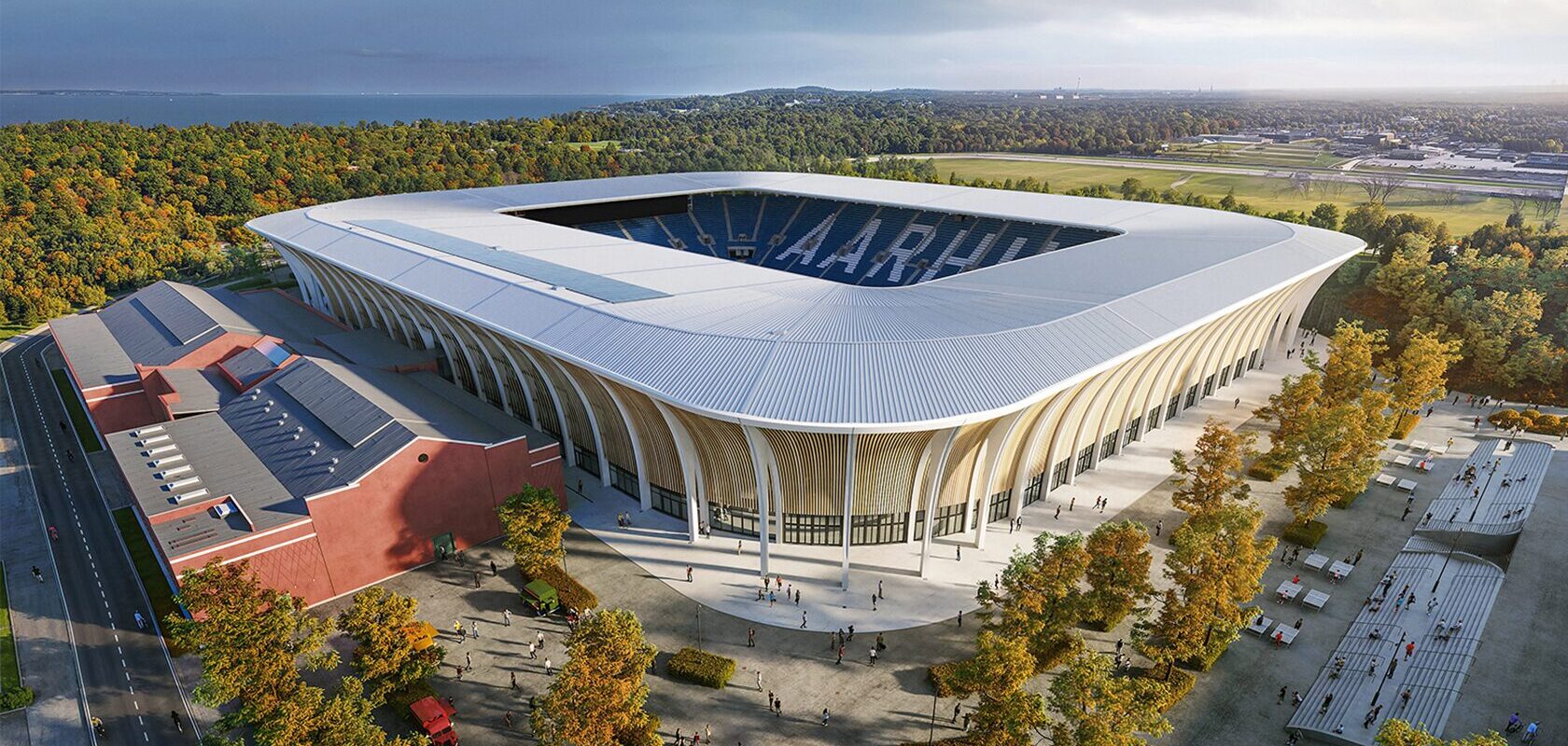 Luftfoto af moderne stadion i Aarhus, omgivet af skov og bygning med rødt tag.