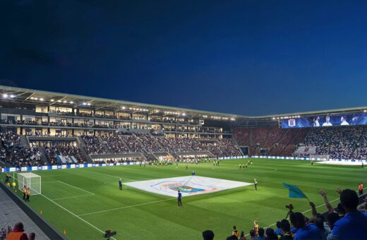 Fodboldstadion om aftenen med fans, spillere og et stort banner på banen under kampforberedelse.