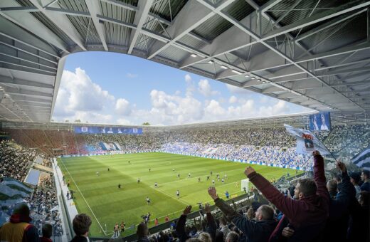 Fyldt fodboldstadion under kamp, fans jubler, blå himmel, stort udendørs sportsarrangement.