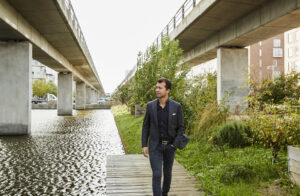 Dariush Rezai går langs kanal under viadukt, iført jakkesæt.
