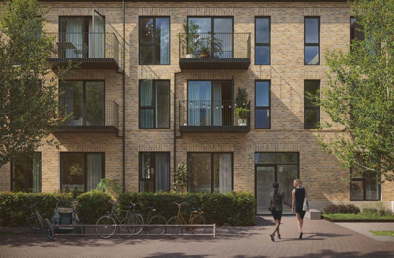 Murstensbygning med altaner, cykler og to personer som går mod indgangen. Grønt buskads foran bygningen.