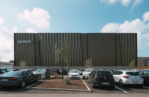 Norlys bygning med parkeringsplads og biler under blå himmel og skyer, set udefra.