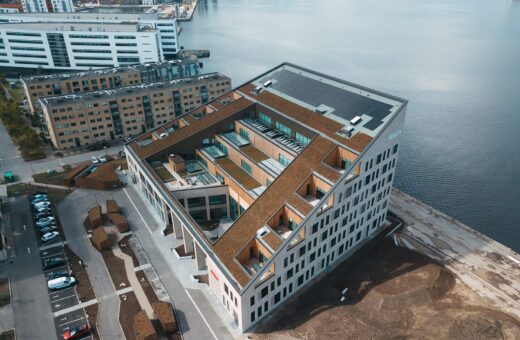 Luftfoto af moderne, stor bygning med skrå tag, beliggende ved vandet og omgivet af andre beboelsesbygninger.