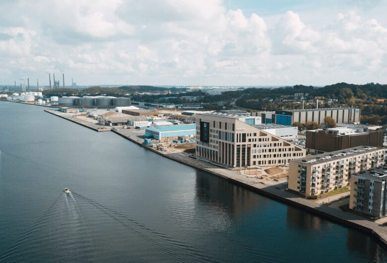 Luftfoto af en havnefront med bygninger, lagerhuse og to både på vandet under en delvist skyet himmel.