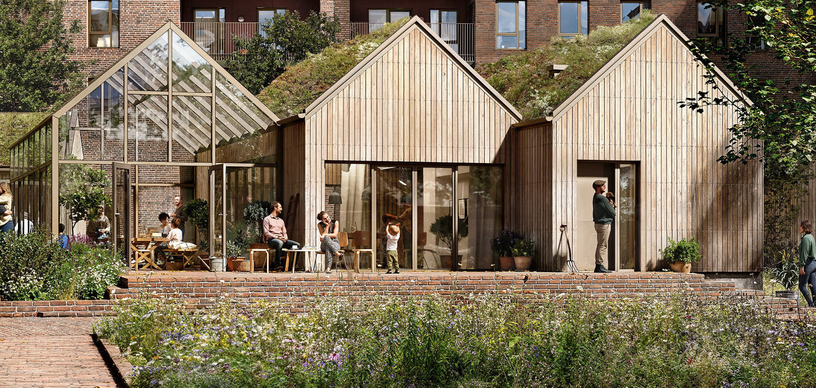 Moderne nybyggeri med træhuse og drivhus, mennesker sidder og står udendørs, blomsterhave foran.