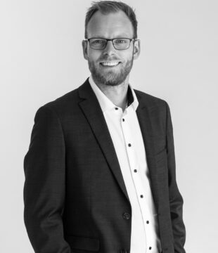 Mand med briller i hvid skjorte og mørk blazer på grå baggrund i sort/hvid