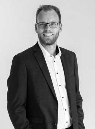 Mand med briller i hvid skjorte og mørk blazer på grå baggrund i sort/hvid