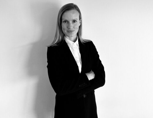 Person står med korslagte arme iført en mørk jakke og hvid skjorte mod en hvid baggrund.