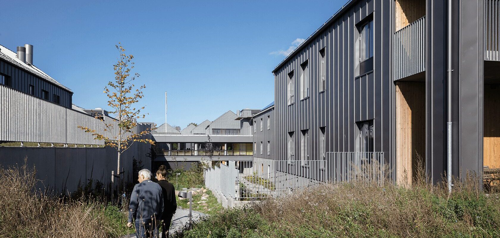 Moderne bygninger med grå facader, to personer går på en sti, blå himmel.