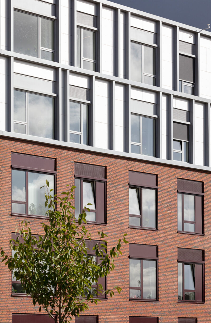 Moderne bygning med murstens- og metaldetaljer, store vinduer og et træ i forgrunden.