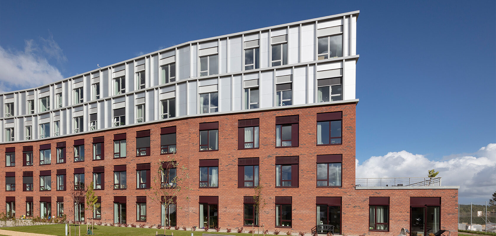 Moderne bygning med røde mursten og hvide paneler under en blå himmel. Person med rollator foran.