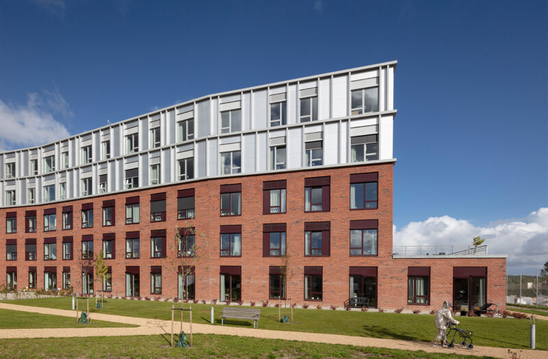 Moderne bygning med røde mursten og hvide paneler under en blå himmel. Person med rollator foran.