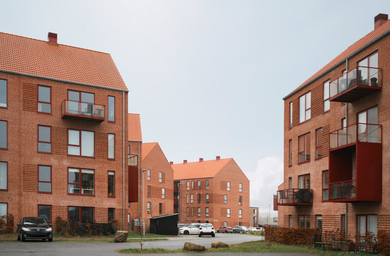 Røde murstensbygninger med altaner, parkeringsområde og biler i en moderne boligkompleks.