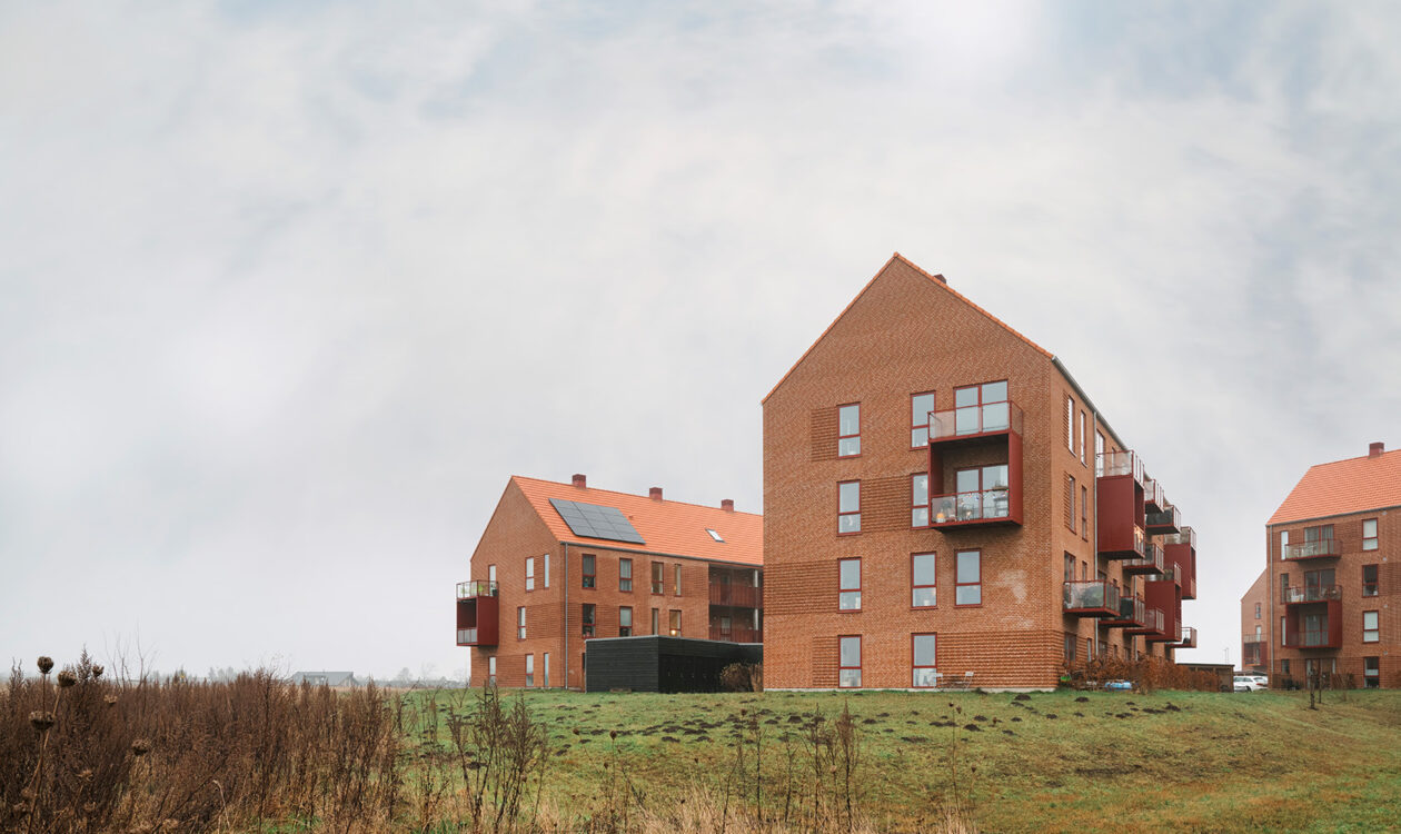 Tre moderne murstensbygninger med rødt tag, græsareal i forgrunden og overskyet himmel.