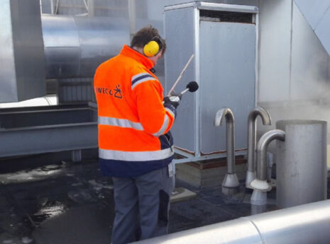 Person måler støjniveau ved ventilationsanlæg iført orange jakke og høreværn.