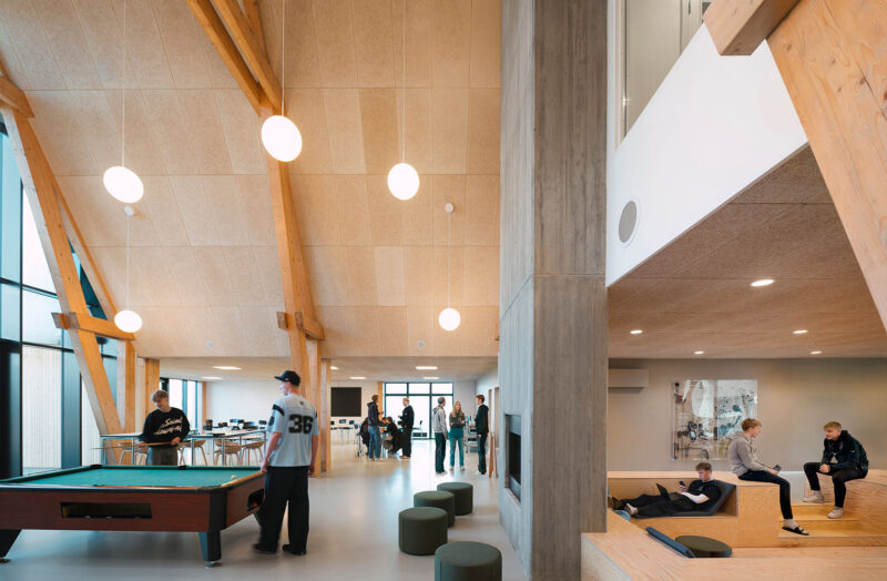 Moderne fællesrum med billardbord, høj loftshøjde, træ- og betonindretning, personer socialiserer.