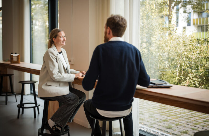 To kolleger holder møde ved højt bord i lyst kontor med store vinduer