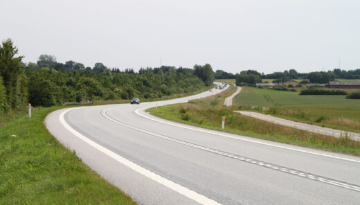 Snoet landevej gennem grønt dansk landskab med få biler og cykelsti ved siden af