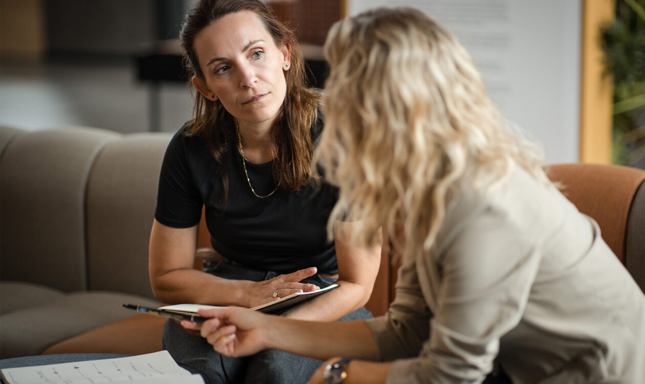To kvinder i samtale på sofa med notesbøger under et uformelt møde