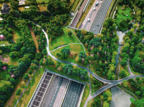 Luftfoto af motorvej med grøn park, stier og broer omgivet af træer og huse