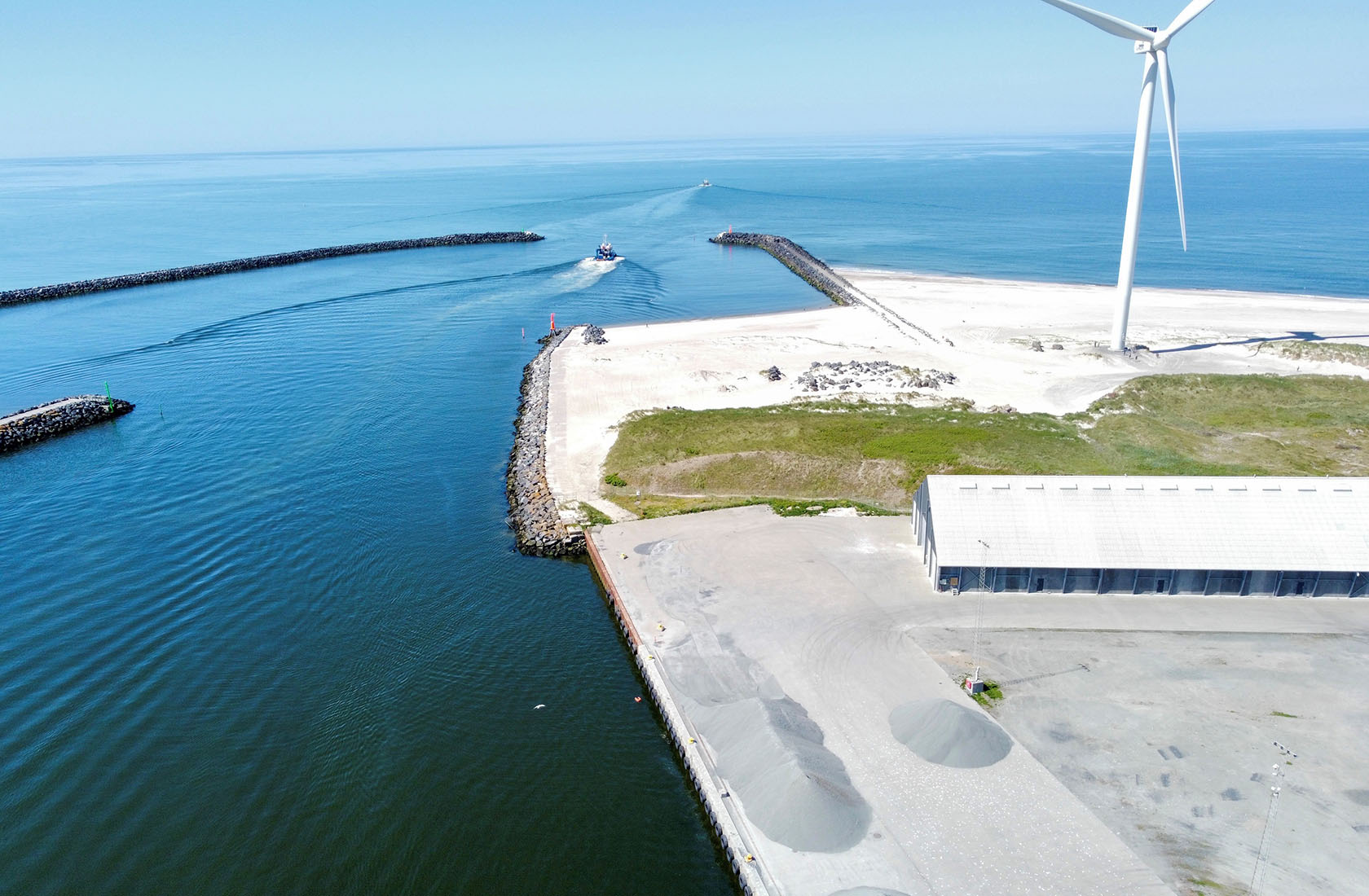 Luftfoto af havneindsejling med vindmølle, kajanlæg og sejlbåd på blåt hav
