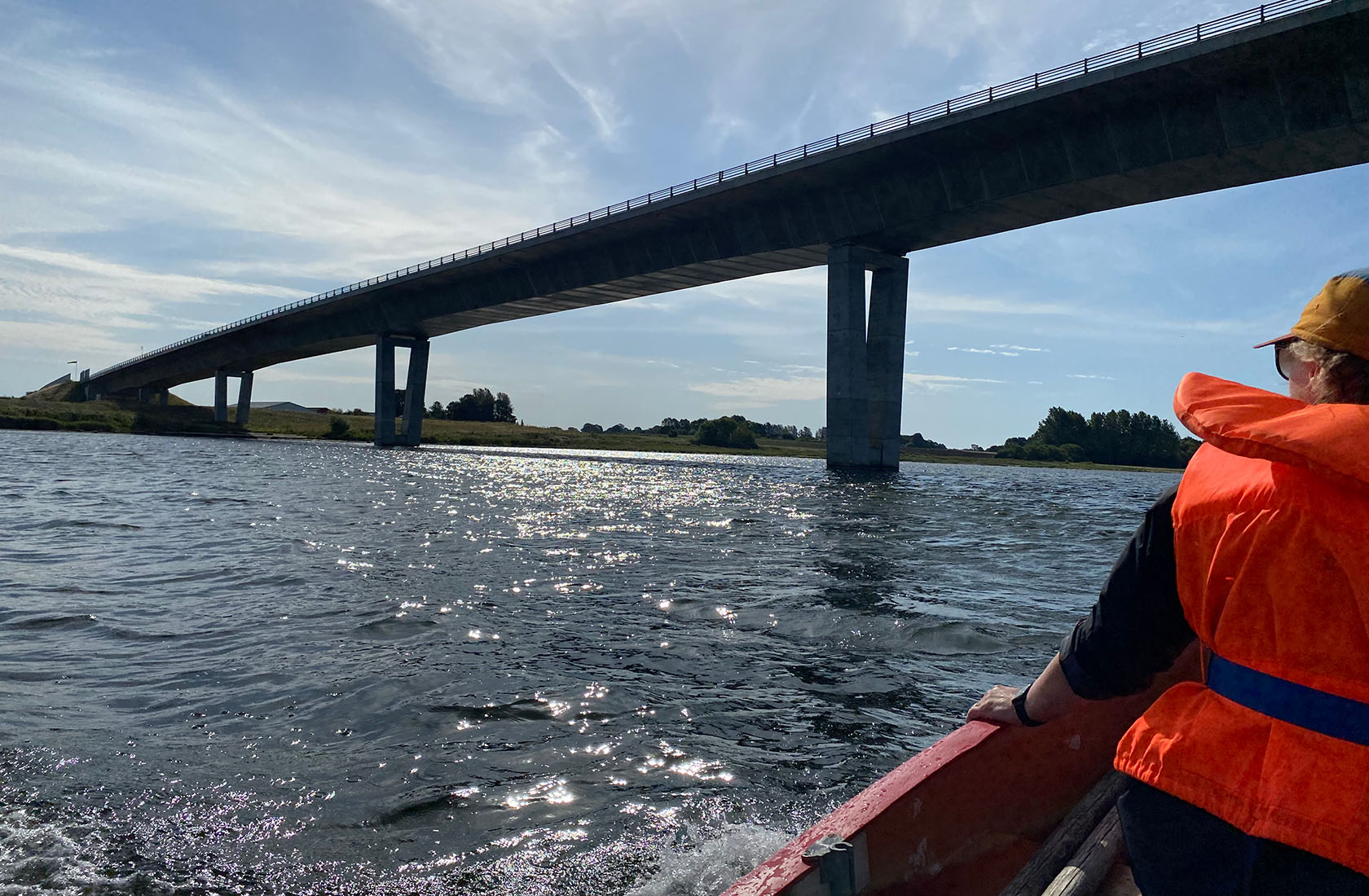 Person i orange redningsvest sejler under stor bro på solbeskinnet fjord